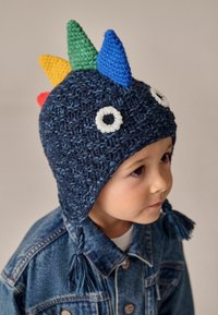 Gehaakte marineblauwe muts met kleurrijke pieken (groen, blauw, geel, rood) en witte ogen. Bovenop de oorflappen hangen franjes, die zowel warmte als stijl bieden.