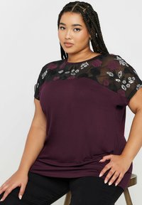 Marooni yläosa, jossa on läpikuultava musta kukkakirjailtu verkko-osa, lyhyet hihat ja rento istuvuus. Pehmeä kangas, jossa on hienovarainen rakenne.