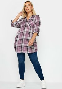 Tunic a scacchi viola e bianchi con scollo a V, maniche lunghe e tasca sul petto. Abbinata a jeans skinny blu scuro e sneaker bianche.