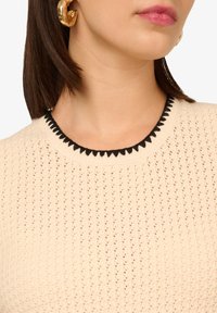 Beige Strickpullover mit einem strukturierten Muster. Schwarzer gewellter Saum am Halsauschnitt. Große goldene Creole sichtbar.