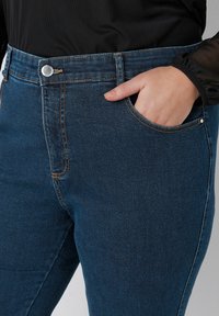 Tummansiniset farkut, joissa on sileä pinta, varustettuna etutaskulla ja hopeisella nappikiinnityksellä, jotka ovat täydentäneet ruskeat ompeluseikat.