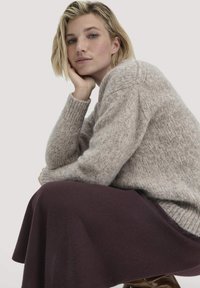 Hellgrauer Wollpullover mit weicher Textur und gerippten Bündchen, kombiniert mit einem fließenden dunkelburgunderfarbenen Rock. Das Model posiert mit einem nachdenklichen Ausdruck.