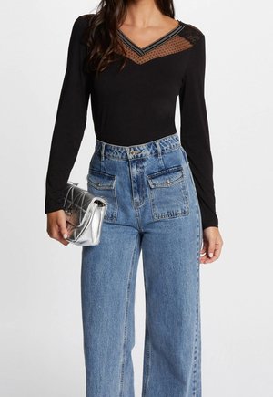 Femme portant un haut noir à manches longues avec un décolleté transparent et un jean bleu taille haute, tenant une petite pochette matelassée argentée.