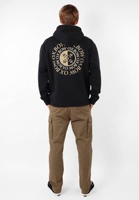 Un sweat à capuche noir avec un grand logo circulaire beige au dos, associé à un pantalon cargo kaki avec des poches latérales et une coupe décontractée.