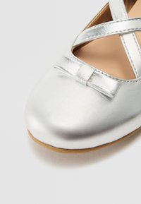 Ballerine metalliche argento con punta rotonda, dotate di un accento a fiocco e di cinturini incrociati. Texture liscia con soletta color beige.