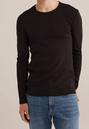 Homme portant une chemise noire unie à manches longues associée à un jean en denim bleu, debout devant un fond neutre clair.