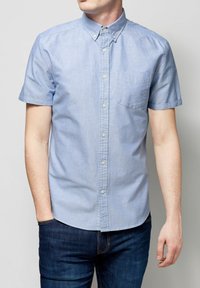 Camisa de botones de manga corta en algodón azul claro con un bolsillo en el pecho y una ligera textura, combinada con jeans de mezclilla azul oscuro.
