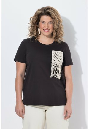 Frau mit lockigem Haar lächelt, trägt ein schwarzes kurzärmeliges Oberteil mit einer weißen Makramee-Tasche und hellfarbige Hosen.