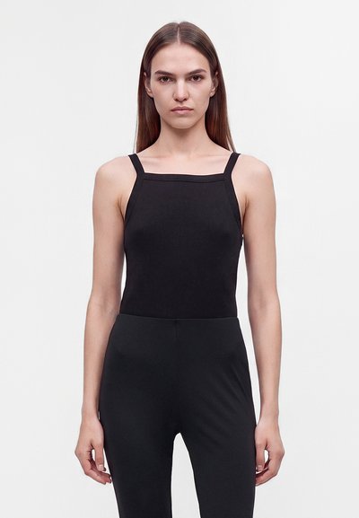 Jeune femme aux longs cheveux bruns portant un haut noir sans manches ajusté et un pantalon noir, debout devant un fond blanc uni.