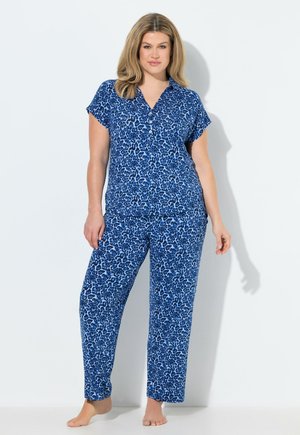 Donna in piedi a piedi nudi che indossa una maglietta a maniche corte con stampa leopardata blu e pantaloni lunghi abbinati su sfondo bianco.