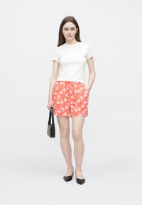 Bílé tričko s kulatým výstřihem spojené s červenými šortkami s květinovým vzorem. Modelka drží malou černou kabelku a má na sobě černé baleríny.