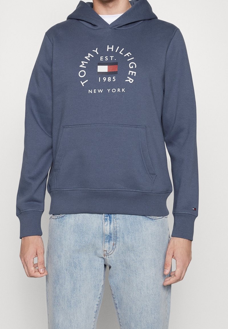 Námořnická modrá mikina s kapucí a přední kapsou, s kulatým logem a textem "TOMMY HILFIGER EST. 1985 NEW YORK" a barevnými akcenty.
