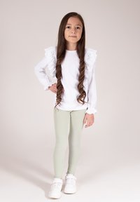 Biała bluzka z długim rękawem ozdobiona falbankami na ramionach, zestawiona z jasnooliwkowymi prążkowanymi legginsami i białymi tenisówkami. Proste brązowe włosy.