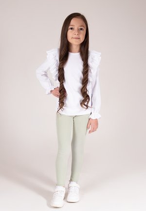 Biała bluzka z długim rękawem ozdobiona falbankami na ramionach, zestawiona z jasnooliwkowymi prążkowanymi legginsami i białymi tenisówkami. Proste brązowe włosy.