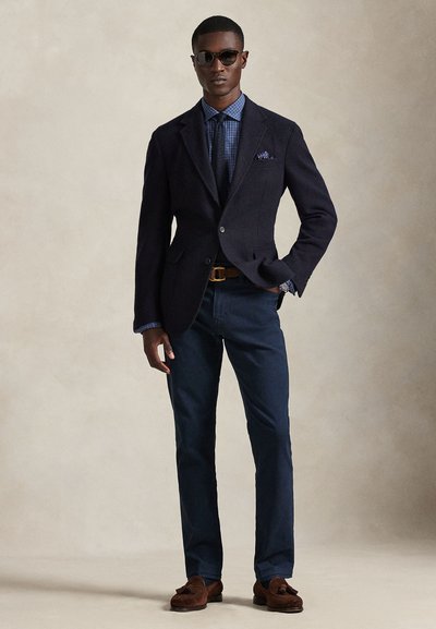 Blazer azul-marinho, camisa azul com padrão, calças escuras, cinto castanho e mocassins. Óculos de sol completam o visual. Texturas suaves e corte ajustado.