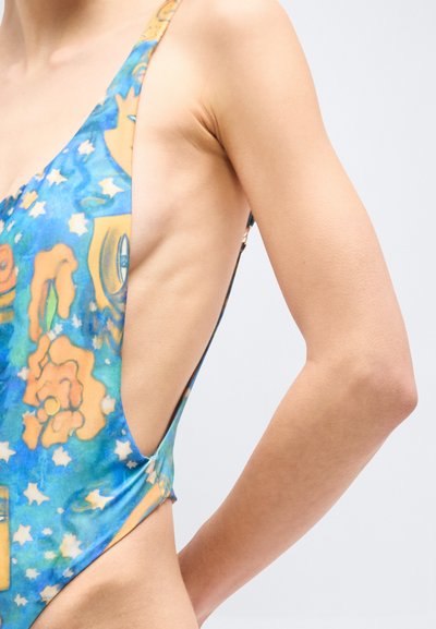 Πλάγια όψη ατόμου που φοράει μαγιό με μπλε και πορτοκαλί μοτίβα και βαθύ κόψιμο κάτω από τη μασχάλη, σε απλό φόντο.