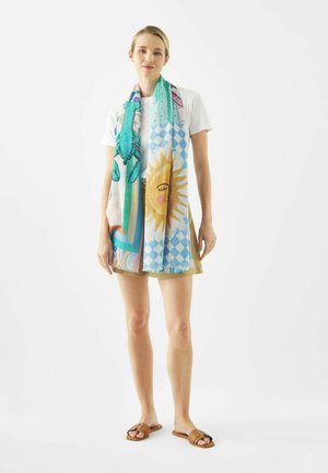 Vrouw draagt een wit T-shirt, een kaki rok, sandalen en een kleurrijke sjaal met zon- en abstracte patronen, staand tegen een witte achtergrond.