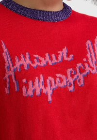 Red knitted sweater featuring a purple, sparkly collar and textured design with the words “Amour impossibil” in blue and pink lettering.