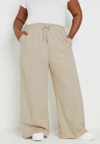 Beige levelekkaiset housut, joissa on joustava naruhame, etutaskut ja sileä rakenne, paritettu valkoisten lenkkareiden kanssa.