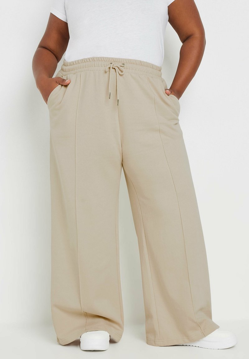 Beige levelekkaiset housut, joissa on joustava naruhame, etutaskut ja sileä rakenne, paritettu valkoisten lenkkareiden kanssa.