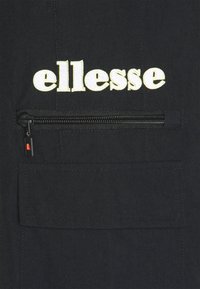 Svart tygjacka med ett broderat "ellesse"-logotyp i vitt, en zipficka och en texturerad yta med kontrasterande sömmar.