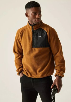 Hombre con un jersey de forro polar marrón con bolsillo negro en el pecho, sosteniendo una botella de agua negra, mirando hacia la izquierda sobre un fondo neutro.