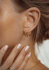 Des boucles d'oreilles en or pendent du lobe de l'oreille. La peau est lisse et douce, avec des ongles fraîchement manucurés présentant une teinte rose et des bords blancs.