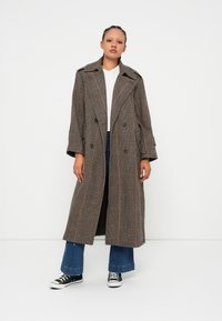 Long, double-breasted coat in a brown and gray plaid pattern. Features a wide collar, buttoned sleeves, and a mid-length design. Paired with flared jeans and sneakers.