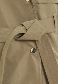 Detailní záběr na pas béžového trenčkotu s prošitou opaskou uvázanou na uzel a lesklým zlatým knoflíkem na látce.