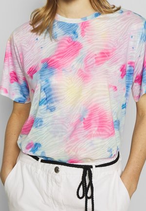 Femme portant un t-shirt à manches courtes tie-dye pastel avec des motifs roses, bleus et jaunes, rentré dans un pantalon blanc avec une ceinture corde noire.
