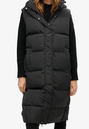 Zwarte lange gewatteerde mouwloze bodywarmer met een rits aan de voorkant en drukknopen, gedragen over een zwarte trui, die de heupen bedekt.