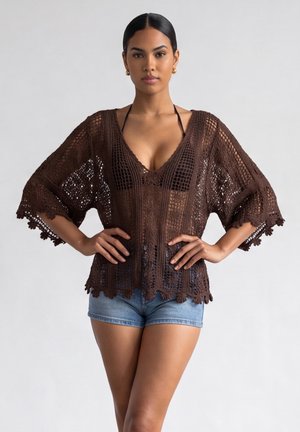 Donna che indossa un top marrone all'uncinetto sopra un bikini nero e pantaloncini di jeans, in piedi con le mani sui fianchi contro uno sfondo semplice.