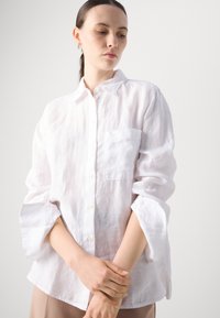 Camicia bianca button-down realizzata in lino leggero con vestibilità comoda, caratterizzata da una tasca sul petto e maniche arrotolate. Texture liscia.