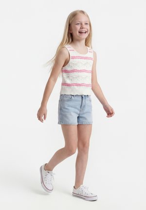 Mladé dievča kráča, oblečené v bielej pletenej tielkovej blúzke s ružovými pruhmi, svetlomodrých rifľových šortkách a bielych teniskách.