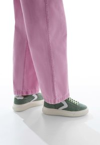 Jeans rosa a gamba larga con una texture liscia e cuciture visibili, abbinati a sneaker basse in suede verde con suola in gomma e accenti bianchi.