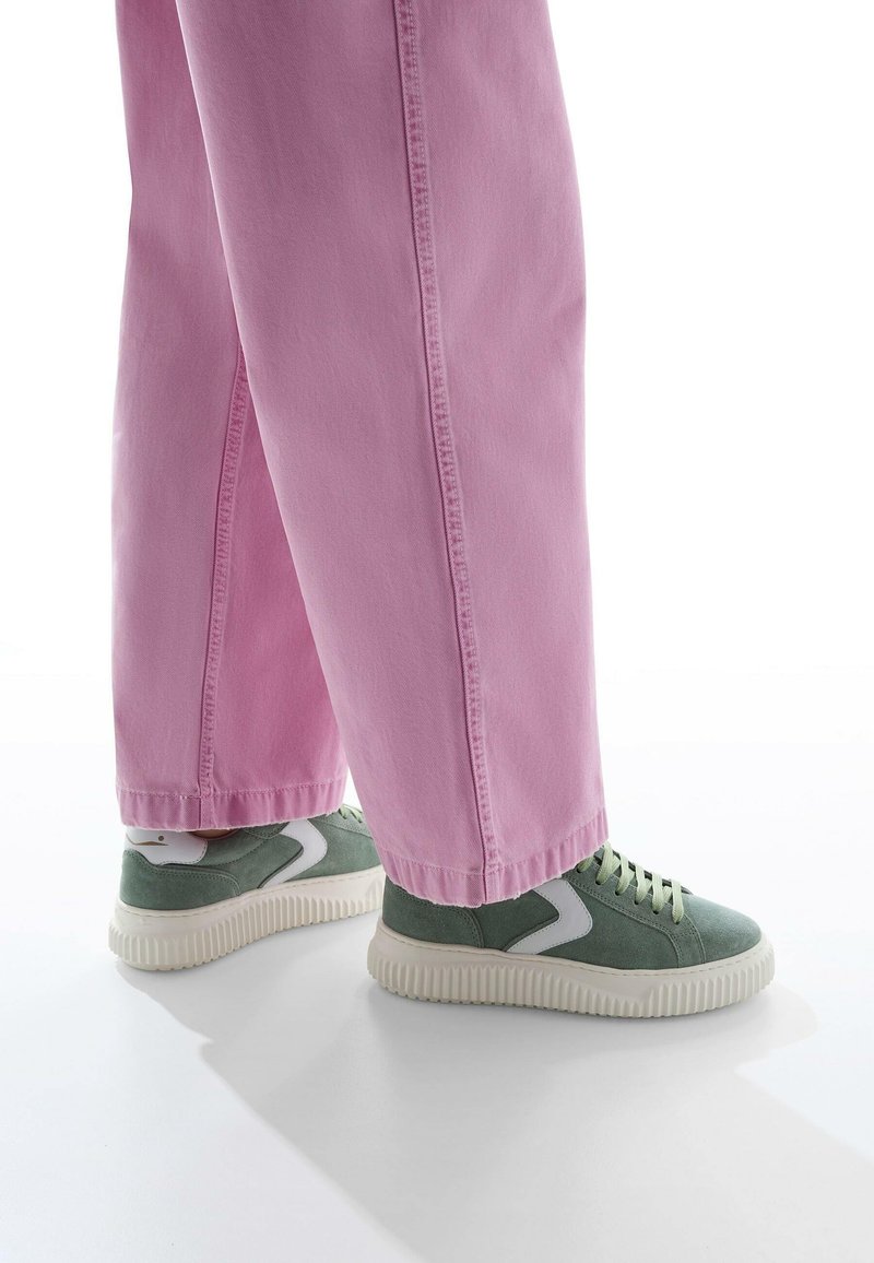 Jeans rosa a gamba larga con una texture liscia e cuciture visibili, abbinati a sneaker basse in suede verde con suola in gomma e accenti bianchi.