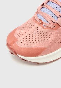 Lys pink atletisk sko med et meshned, perforeringer for åndbarhed og en hvid mellemsål. Har flade snørebånd og en rund tå.