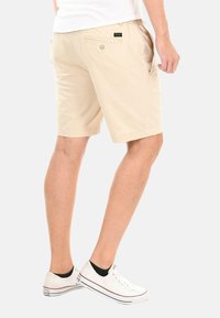 Beige Shorts aus leichtem Stoff, mit seitlichen und hinteren Taschen sowie einer lässigen Passform, kombiniert mit weißen Sneakern.