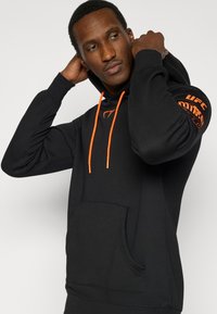 Μαύρο φούτερ με πορτοκαλί κορδόνια και λεπτομέρειες. Διαθέτει μπροστινή τσέπη και το λογότυπο "UFC" τυπωμένο στο αριστερό μανίκι. Μαλακή υφή υφάσματος.