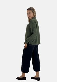Groene langemouwen shirt met een gestructureerd oppervlak, gecombineerd met zwarte cropped wijde jeans en platte schoenen, zijdelings getoond.