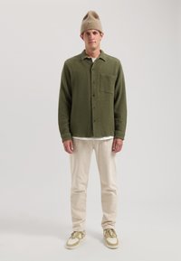 Jachetă cămașă texturată verde, cu închidere cu nasturi și un buzunar pe piept, asortată cu pantaloni din bumbac crem și adidași măslinii.