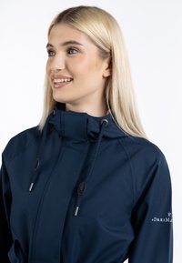 Jachetă impermeabilă de culoare albastru închis, cu guler înalt, glugă cu cordon și accente de metal argintiu. Include logo-ul pe mânecă. Textură netedă.