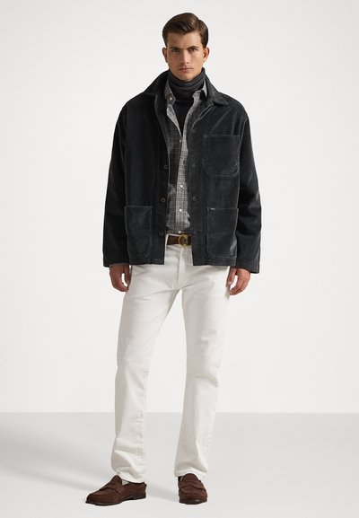 Black corduroy jacket with two front pockets, worn over a gray and white checked shirt. White trousers and brown loafers complete the outfit.