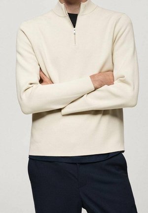 Personne les bras croisés portant un pull crème à demi-fermeture éclair sur une chemise noire et un pantalon sombre sur un fond uni.