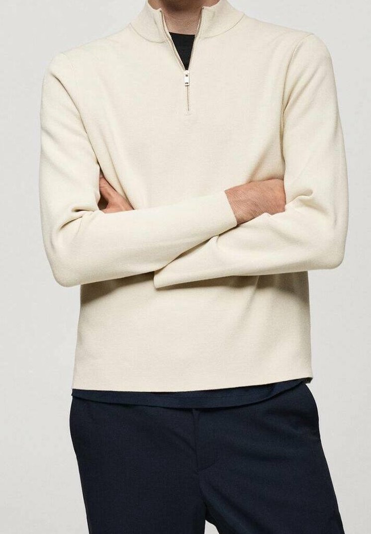 Personne les bras croisés portant un pull crème à demi-fermeture éclair sur une chemise noire et un pantalon sombre sur un fond uni.