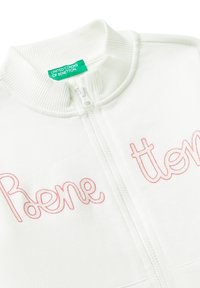 Biała kurtka zapinana na zamek z prążkowanym kołnierzem. Zawiera różowy haftowany napis "Roene." Metka widoczna w okolicy szyi. Miękka tekstura materiału.