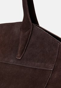 Σκούρο καφέ σουέτ τσάντα χειρός με δομημένο σχήμα, που διαθέτει έντονα ραψίματα και επίπεδο χερούλι για εύκολη μεταφορά.