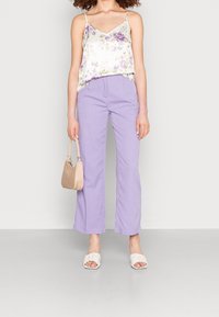 Femme portant un pantalon taille haute lilas, un haut sans manches blanc à fleurs, des talons ouverts blancs, et tenant un sac à main beige.