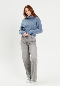 Light blue turtleneck sweater, soft knit texture, paired with high-waisted grey jeans and multi-colored sneakers. Model stands smiling.