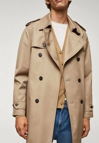 Beige trenchcoat met dubbele rij knopen, zwarte knopen en schouderflappen. Draag het over een beige cardigan en een wit t-shirt.